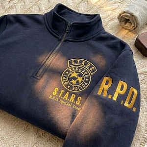 Könnte beinhalten: Marineblaues Sweatshirt mit einem Kragen mit Viertelreißverschluss und goldfarben gesticktem Text und Aufnähern. Die Aufnäher lauten "S.T.A.R.S. Raccoon Police Dept." und "R.P.D. Special Team". Das Sweatshirt hat ein verwaschenes, verblichenes Aussehen.