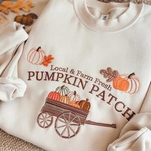 Può includere: Felpa color crema con ricami di zucche, foglie e un carretto. Il testo "Local & Farm Fresh PUMPKIN PATCH" è ricamato in marrone. Il design presenta varie zucche nei toni dell'arancione, verde e marrone.