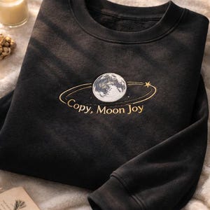 Puede incluir: Sudadera negra de cuello redondo con un diseño bordado que presenta un globo terráqueo, un anillo dorado y una estrella. El texto "Copy, Moon Joy" está bordado en dorado. La sudadera está doblada sobre una superficie suave y texturizada.