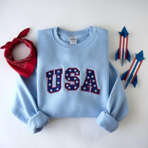 May include: A pale blue sweatshirt featuring the word "USA" in red and blue, star-patterned lettering. A red bandana and two paper rockets in red, white, and blue are also visible. The sweatshirt has a crew neck and long sleeves.