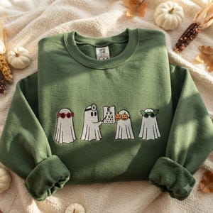 May include: An olive green crewneck sweatshirt featuring an embroidered design of four white ghosts. One ghost wears red glasses, another wears a head mirror and holds an eye chart, one wears pumpkin glasses, and the last wears green bat glasses.