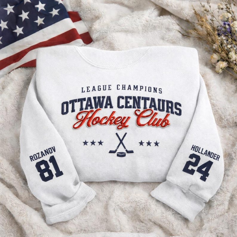 May include: White sweatshirt with the text "LEAGUE CHAMPIONS OTTAWA CENTAURS Hockey Club". The sleeves display the numbers "81" and "24" with player names. An American flag is in the background.