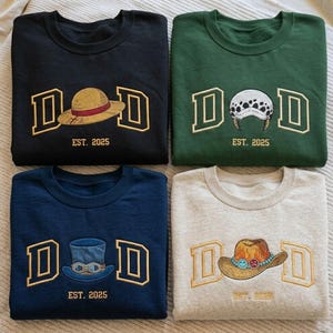 Könnte beinhalten: Vier Sweatshirts mit Rundhalsausschnitt in Schwarz, Grün, Marineblau und Grau. Jedes zeigt das gestickte Wort "DAD" in goldenen Blockbuchstaben mit einem Hutdesign in der Mitte und "EST. 2025" darunter. Die Hüte variieren in Stil und Farbe.