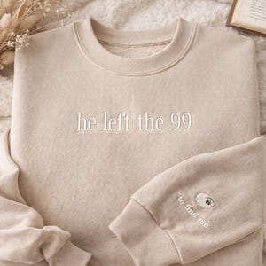 May include: A beige sweatshirt with the embroidered text "he left the 99" across the chest. The sleeve cuff features a sheep graphic and the text "to find me". The sweatshirt is made of a soft material.