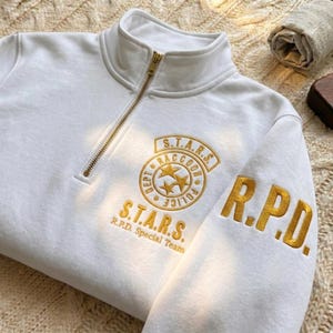 May include: A white quarter-zip sweatshirt featuring gold embroidered text and graphics. The chest displays a circular S.T.A.R.S. Raccoon Police Dept emblem with three stars, while the sleeve features the bold gold embroidered letters R.P.D.