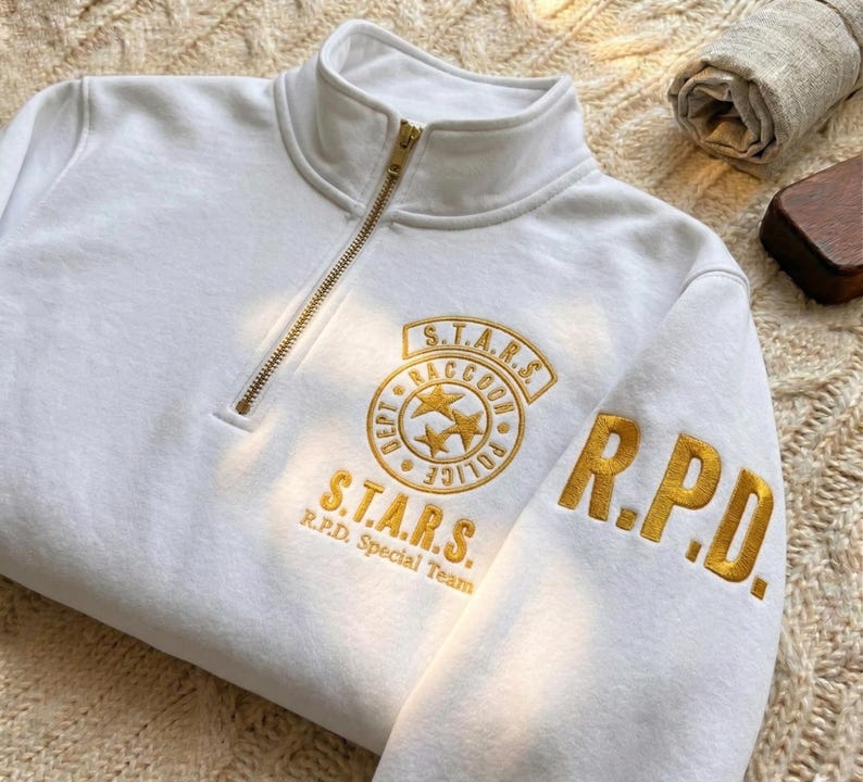 May include: A white quarter-zip sweatshirt featuring gold embroidered text and graphics. The chest displays a circular S.T.A.R.S. Raccoon Police Dept emblem with three stars, while the sleeve features the bold gold embroidered letters R.P.D.