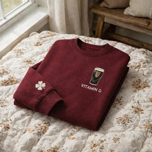 May include: A maroon sweatshirt with a crew neck, featuring a white and black graphic of a pint of stout and the text "VITAMIN G." The left sleeve cuff has a white embroidered four-leaf clover.