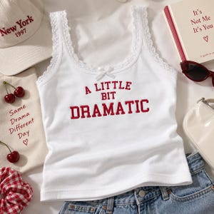 May include: A white sleeveless tank top with lace trim along the neckline and straps, featuring a small white bow at the center. The text A LITTLE BIT DRAMATIC is embroidered in red letters on the front of the garment, which is made of fabric.