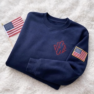 Könnte beinhalten: Marineblaues Sweatshirt mit rotem, gesticktem Logo und einem amerikanischen Flaggenaufnäher auf dem Ärmel. Eine kleine amerikanische Flagge liegt neben dem Sweatshirt auf einer weißen, flauschigen Oberfläche.
