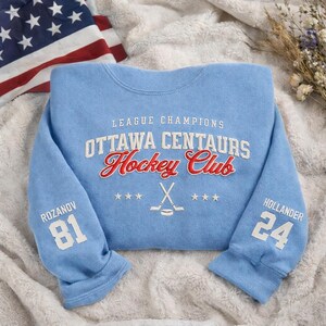 May include: Light blue Ottawa Centaurs Hockey Club sweatshirt. The text "League Champions" and "Ottawa Centaurs Hockey Club" are in white and red. The sleeves have the names and numbers "Rozanov 81" and "Hollander 24". An American flag is in the background.