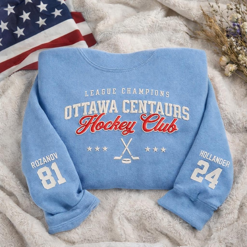 May include: Light blue Ottawa Centaurs Hockey Club sweatshirt. The text "League Champions" and "Ottawa Centaurs Hockey Club" are in white and red. The sleeves have the names and numbers "Rozanov 81" and "Hollander 24". An American flag is in the background.