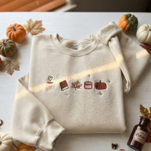 May include: A cream-colored crewneck sweatshirt featuring small, centered embroidery of a coffee mug with a heart, a book, a maple leaf, a candle, and a pumpkin, accented by tiny star shapes. The fabric is a solid, light beige cotton blend.