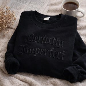Op de afbeelding: Zwart sweatshirt met de geborduurde tekst "Perfectly Imperfect" in een gotisch lettertype. Het kledingstuk wordt getoond op een zacht, getextureerd oppervlak, met een kopje koffie en een krant op de achtergrond.