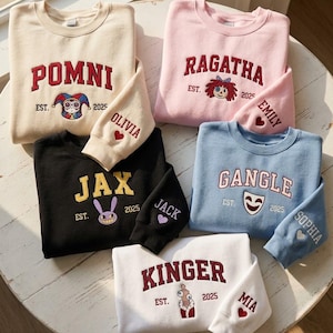 May include: Five crewneck sweatshirts in beige, pink, black, light blue, and white. Each features embroidered character names, EST. 2025, and a character graphic. Sleeves are embroidered with a name and a small heart symbol in red, purple, or white.