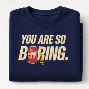 May include: A navy blue sweatshirt with the text "YOU ARE SO BORING." The word "BORING" features a Ginger Ale can graphic. A gold-coloured, stylised anchor is next to the can.