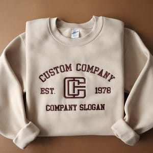 May include: A cream-coloured sweatshirt featuring maroon embroidered text: "CUSTOM COMPANY", "EST. 1978", and "COMPANY SLOGAN". A maroon "C" logo is centered. The sleeves are cuffed. The fabric has a soft appearance.