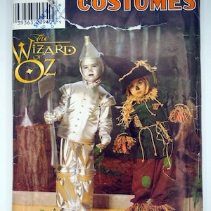 May include: A Simplicity Costumes sewing pattern envelope for The Wizard of Oz featuring a metallic silver Tin Man costume with an axe and a dark green and brown Scarecrow costume with straw accents. The package includes text and a barcode.