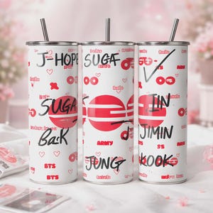 Könnte beinhalten: Drei weiße Edelstahlbecher mit schwarzem Text und roten Akzenten. Jeder Becher trägt einen anderen Namen: J-HOPE, SUGA und JIN, zusammen mit anderem Text und Grafiken. Jeder Becher hat einen silbernen Strohhalm.