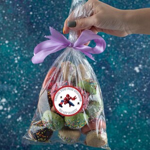 Puede incluir: Una bolsa de pl&aacute;stico transparente llena de galletas coloridas, atada con una cinta morada. Una pegatina con un superh&eacute;roe de dibujos animados y el texto "Gracias por venir a mi fiesta" est&aacute; pegada a la bolsa.