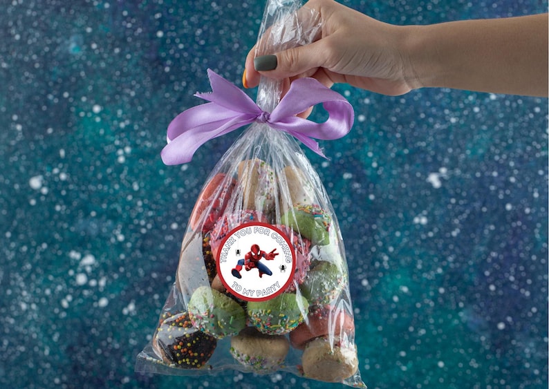 Puede incluir: Una bolsa de pl&aacute;stico transparente llena de galletas coloridas, atada con una cinta morada. Una pegatina con un superh&eacute;roe de dibujos animados y el texto "Gracias por venir a mi fiesta" est&aacute; pegada a la bolsa.