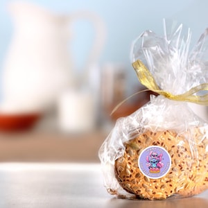 Puede incluir: Dos galletas redondas de color marr&oacute;n claro ligeramente dorado, cubiertas de semillas. Las galletas est&aacute;n envueltas en pl&aacute;stico transparente con una cinta dorada y una pegatina con un personaje de dibujos animados. La pegatina dice "Ohana means family".