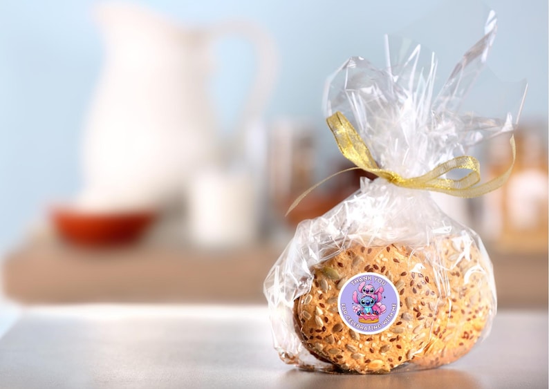 Puede incluir: Dos galletas redondas de color marr&oacute;n claro ligeramente dorado, cubiertas de semillas. Las galletas est&aacute;n envueltas en pl&aacute;stico transparente con una cinta dorada y una pegatina con un personaje de dibujos animados. La pegatina dice "Ohana means family".