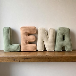 May include: Four plush, three-dimensional letters spelling LENA. The L is sage green, the E is peach, the N is cream, and the A is sage green. Each letter features a consistent, textured, crocheted-style knit surface and rounded edges.