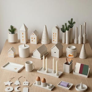 May include: A collection of off-white ceramic home decor items including house-shaped lanterns, conical trees, candle holders, trays, a mug, a pencil cup, egg cups, hanging ornaments, and a soap dish, some featuring small cutouts and lit candles.