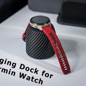 May include: A black, spiralled, cylindrical charging dock for a Garmin watch, featuring a gold-toned watch casing and a vibrant red silicone strap. The text "Charging Dock for Garmin Watch" is printed in black on the white surface below.