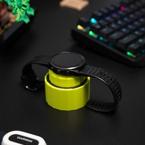 Könnte beinhalten: Eine schwarze Smartwatch mit rundem Zifferblatt und schwarzem Armband liegt auf einem leuchtend gelben Ladeständer. Ein schwarzer Herzfrequenzmesser mit dem Wort "GARMIN" ist sichtbar. Der Hintergrund umfasst eine Tastatur und eine kleine grüne Pflanze.
