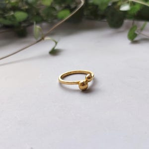 Könnte beinhalten: Ein goldener Ring mit zwei runden, strukturierten Kugeln, die an der Band befestigt sind.