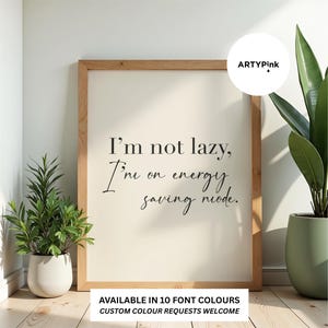 May include: A rectangular wooden frame containing a white print with the black text: I'm not lazy, I'm on energy saving mode. The frame is positioned between two green potted plants. A circular logo in the top right corner reads ARTYPink.