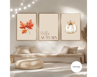 May include: A set of three framed wall art prints featuring a beige background. The left print shows orange maple leaves, the centre print displays the text Hello AUTUMN in dark brown, and the right print depicts a white pumpkin with a bow.