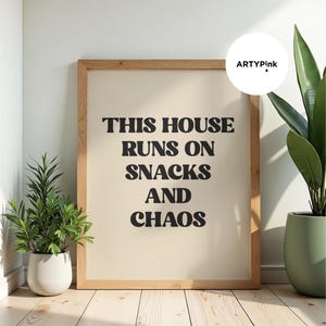 May include: A rectangular wooden frame containing a beige print with the black text THIS HOUSE RUNS ON SNACKS AND CHAOS. The frame is positioned between two potted green plants on a light-colored wooden floor against a white wall.