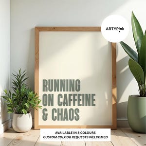 May include: A rectangular wall art print in a light wood frame featuring the text RUNNING ON CAFFEINE & CHAOS in sage green letters on a cream background. The print includes the logo ARTYPink and text stating available in 8 colours.
