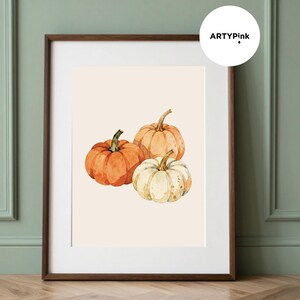 May include: A framed wall art print featuring a watercolour illustration of three pumpkins in shades of orange, light peach, and cream. The artwork is set against a light beige background and enclosed in a dark brown wooden frame with a white mount.