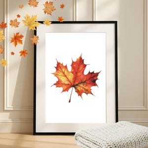 May include: A framed print with a watercolour painting of an autumn maple leaf. The leaf is painted in red, orange, and yellow hues. Decorative maple leaf cutouts are placed around the frame. A folded white blanket rests below.