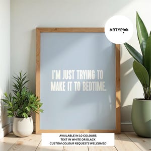 May include: A light blue rectangular wall art print in a wooden frame featuring the white text I'M JUST TRYING TO MAKE IT TO BEDTIME. A circular logo in the top right corner reads ARTYPink. The print is displayed with two potted plants.