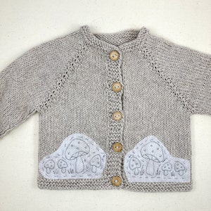May include: A gray knitted cardigan with a button closure and two embroidered mushroom patches on the front.