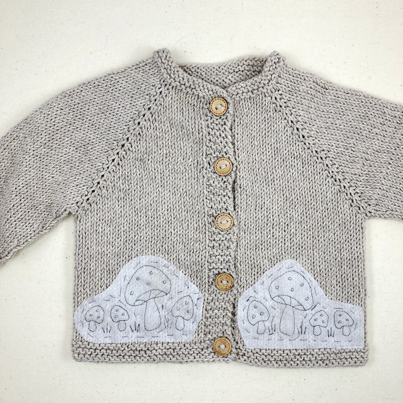 May include: A gray knitted cardigan with a button closure and two embroidered mushroom patches on the front.
