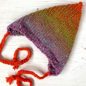Könnte beinhalten: Eine bunte Strickmütze mit einer langen orangenen Schleife. Die Mütze ist aus verschiedenen Farbtönen von lila, blau, grün und orangem Garn gefertigt.
