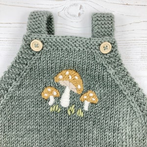 May include: A sage green knitted baby overall with wooden button closures. The front features an embroidered design of three mushrooms with tan caps and white stems. The overall has shoulder straps.