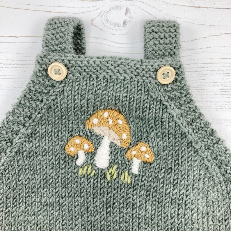 May include: A sage green knitted baby overall with wooden button closures. The front features an embroidered design of three mushrooms with tan caps and white stems. The overall has shoulder straps.