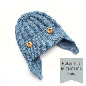 May include: A blue knitted hat with ear flaps and two wooden buttons. The pattern is in English only.