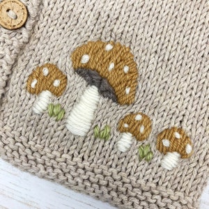 May include: Close-up of a beige knitted fabric with embroidered mushrooms. The mushrooms have brown caps with white spots, white stems, and green grass accents. A wooden button is visible.