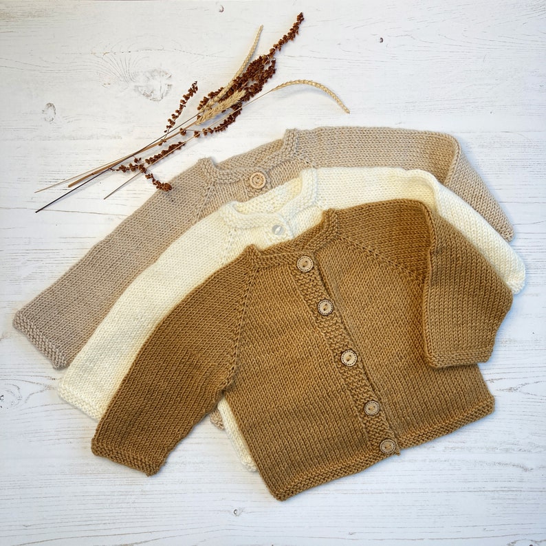 May include: Three long-sleeved knit cardigans in shades of tan, cream, and brown. Each cardigan features a round neckline, ribbed trim, and a front button closure with small wooden buttons. The garments are arranged in a layered, overlapping stack.
