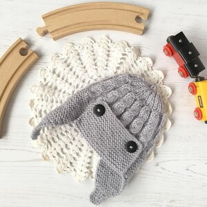 May include: A gray knitted hat with a cable knit pattern and two black buttons. The hat is sitting on a white doily.