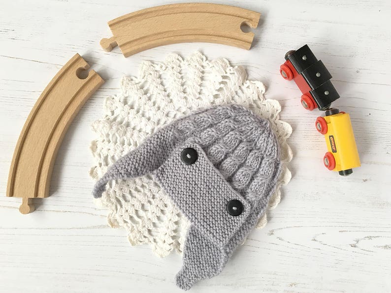 May include: A gray knitted hat with a cable knit pattern and two black buttons. The hat is sitting on a white doily.