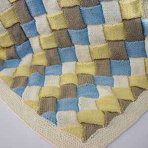 K&ouml;nnte beinhalten: Eine gestrickte Babydecke mit einem gewebten Muster in Blaut&ouml;nen, Gelb und Braun. Die Decke hat einen wei&szlig;en Rand.