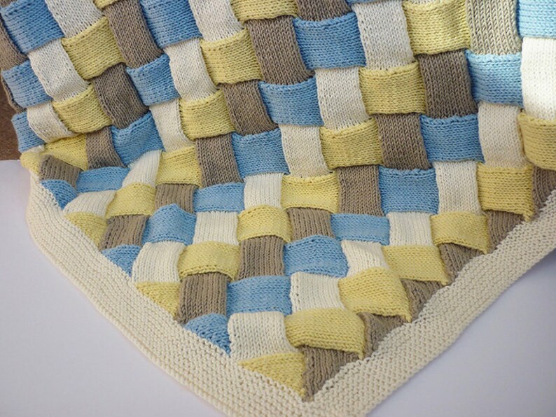 K&ouml;nnte beinhalten: Eine gestrickte Babydecke mit einem gewebten Muster in Blaut&ouml;nen, Gelb und Braun. Die Decke hat einen wei&szlig;en Rand.